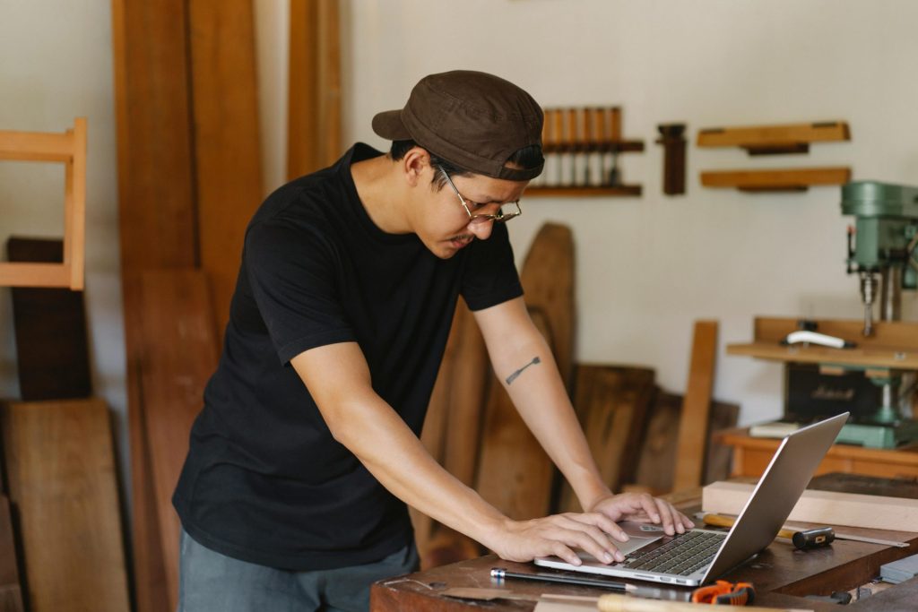 A craftsman focused on a project using a laptop in a woodworking workshop.