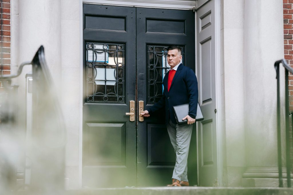 A fashionable man in business attire stands confidently by a building's entrance holding a laptop.