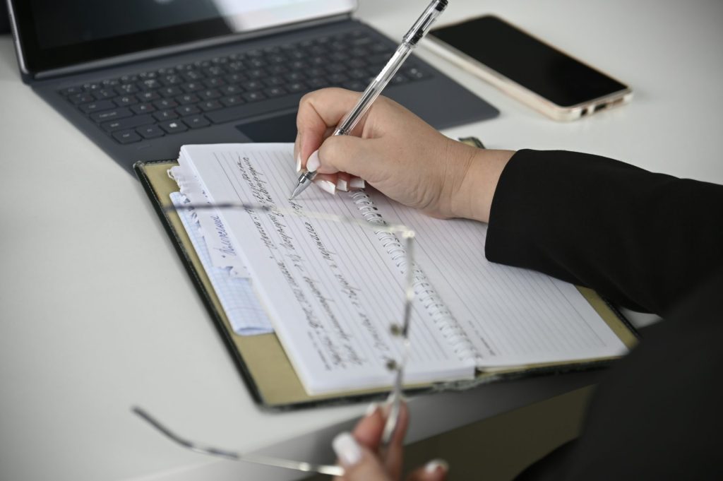 Une personne écrivant sur un cahier avec un ordinateur portable en arrière-plan